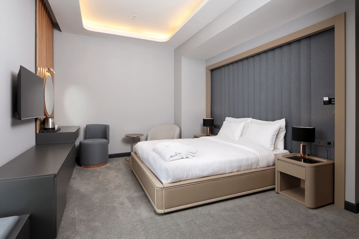 A modern hotel room with a large bed, bedside tables, a flat-screen TV, a desk, and a comfortable armchair.