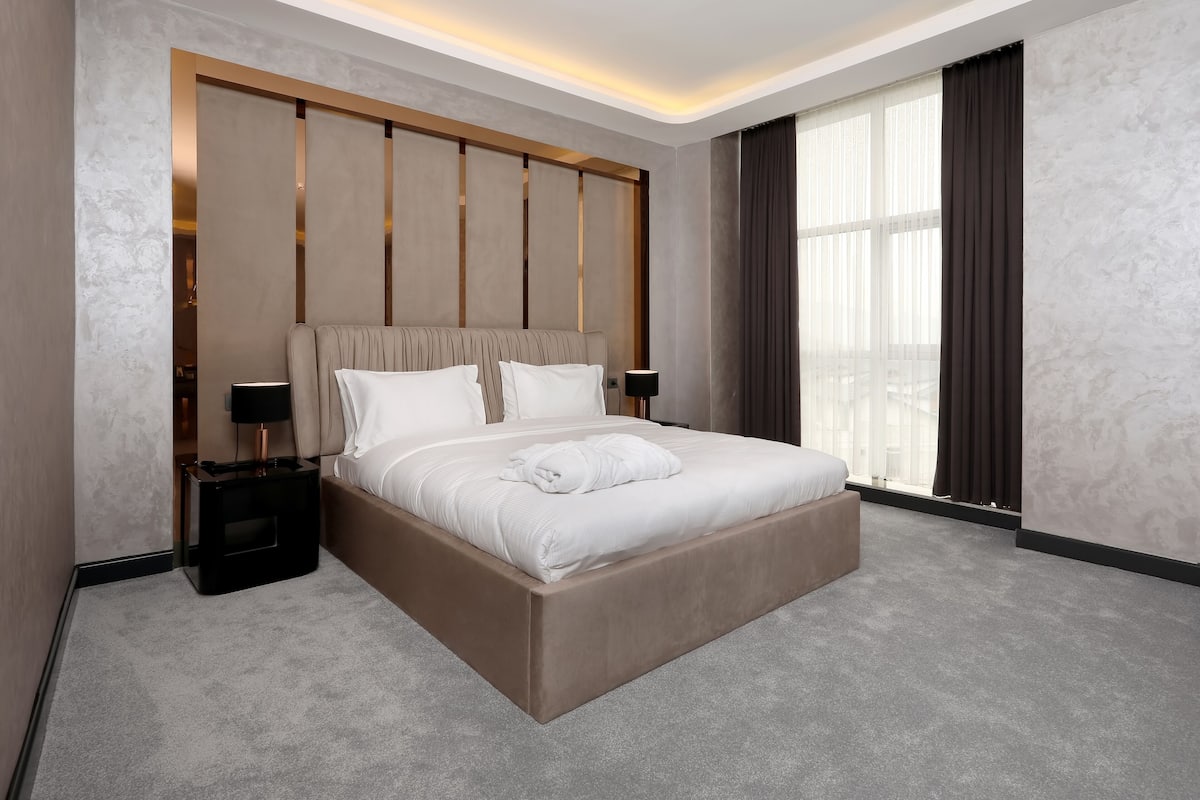 A modern hotel room with a large bed, two bedside tables, a nightstand, and a large window with curtains.
