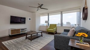 A modern living room with a flat-screen TV, a ceiling fan, a sofa, a coffee table, and a large window with a city view. - 16th FL Bold CozySuites w/pool, gym, roof #5 (Dallas)