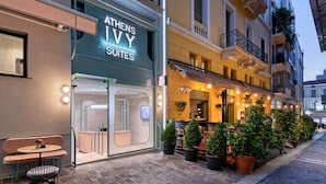 Un hôtel appelé « Athens Ivy Suites », doté d’une façade moderne, d’un espace extérieur aménagé pour s’asseoir et de plantes en pot le long du trottoir.