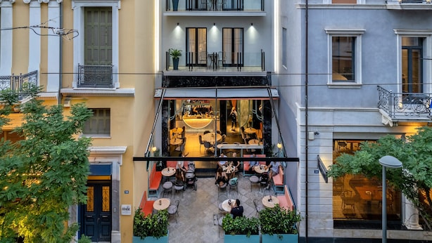 Un immeuble à plusieurs étages doté de balcons, un café situé au rez-de-chaussée avec une terrasse, et un mélange de styles architecturaux.