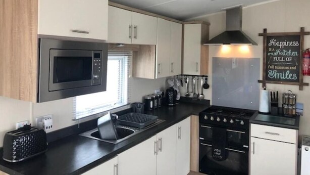 A compact kitchen with a microwave, stove, and sink. There is a chalkboard with a message, and a window providing natural light. - Lovely Holiday Home - Romney Sands by the Sea (Greatstone, New Romney)