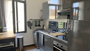 A compact kitchen with a stainless steel refrigerator, a built-in oven, and a sink. There is a window providing natural light, and a small trash bin is visible. - Charming Beach House, Sleeps 10, 150m from Shore (Fort-Mahon-Plage)