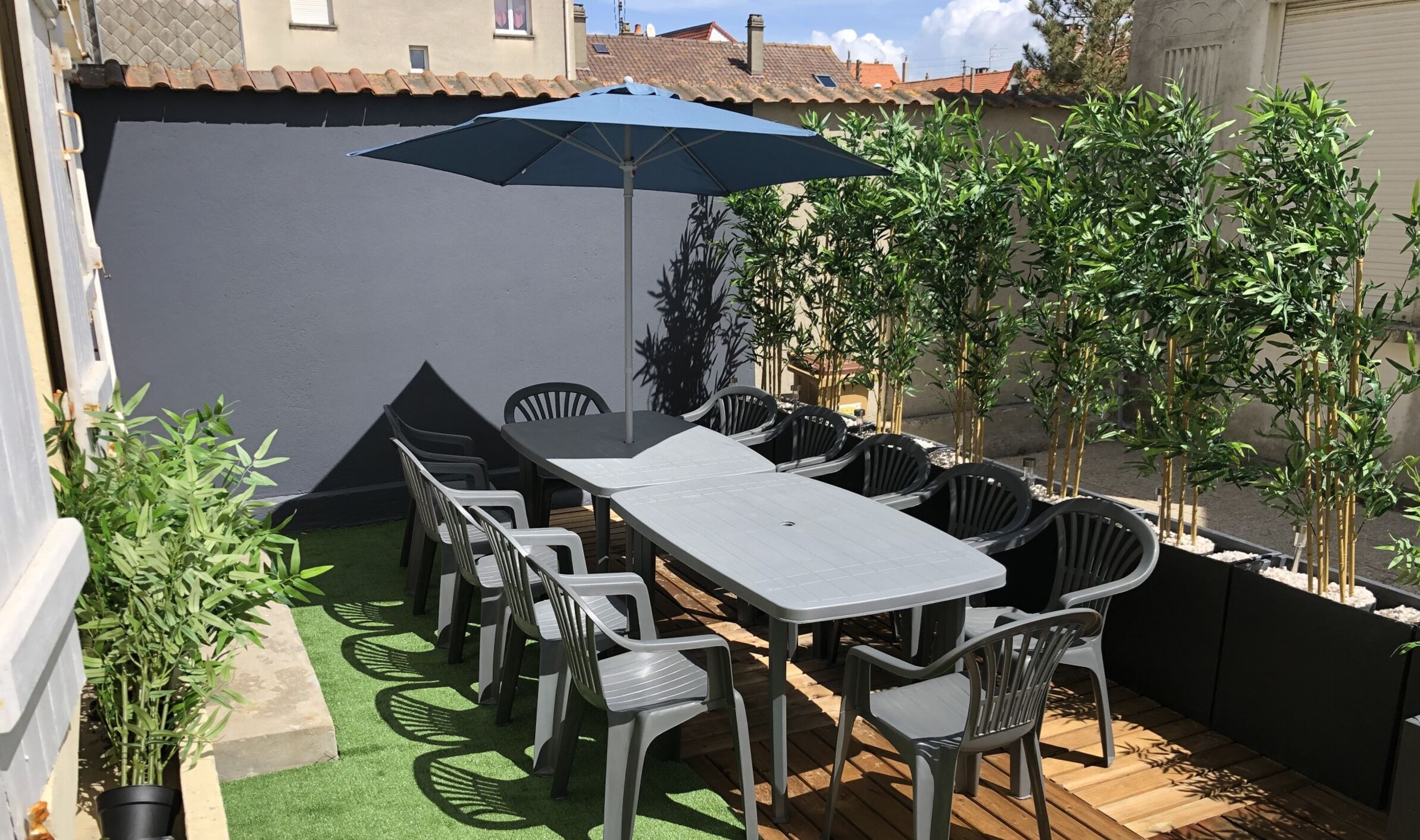 Une terrasse extérieure avec une table et des chaises, entourée de plantes en pot et agrémentée d’un parasol bleu.
