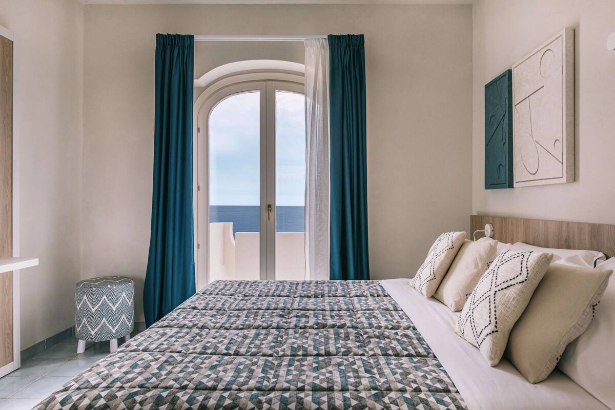 A bedroom with a bed, pillows, a small ottoman, a view through a door, and a wall with a framed picture.
