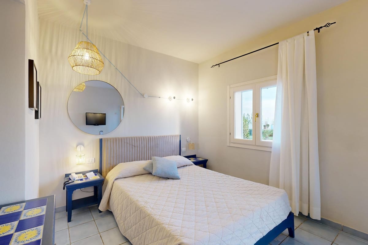 A bedroom with a bed, bedside tables, a TV, a window with curtains, and a wall-mounted mirror.