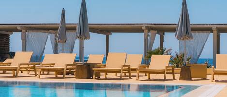 Un espace aménagé au bord de la piscine, avec des chaises longues et des parasols, un ciel dégagé et une vue sur l’océan.