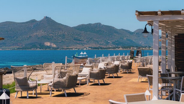 A seaside patio with wicker chairs and tables, overlooking a calm sea and a mountain in the background. - Mangia's Favignana Resort (Favignana)