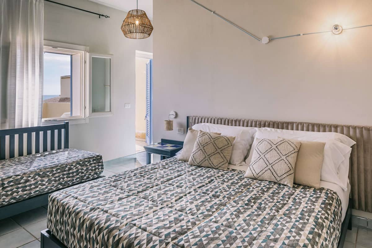 A bedroom with a large bed, two smaller beds, a bedside table, a hanging light fixture, and a window with curtains.