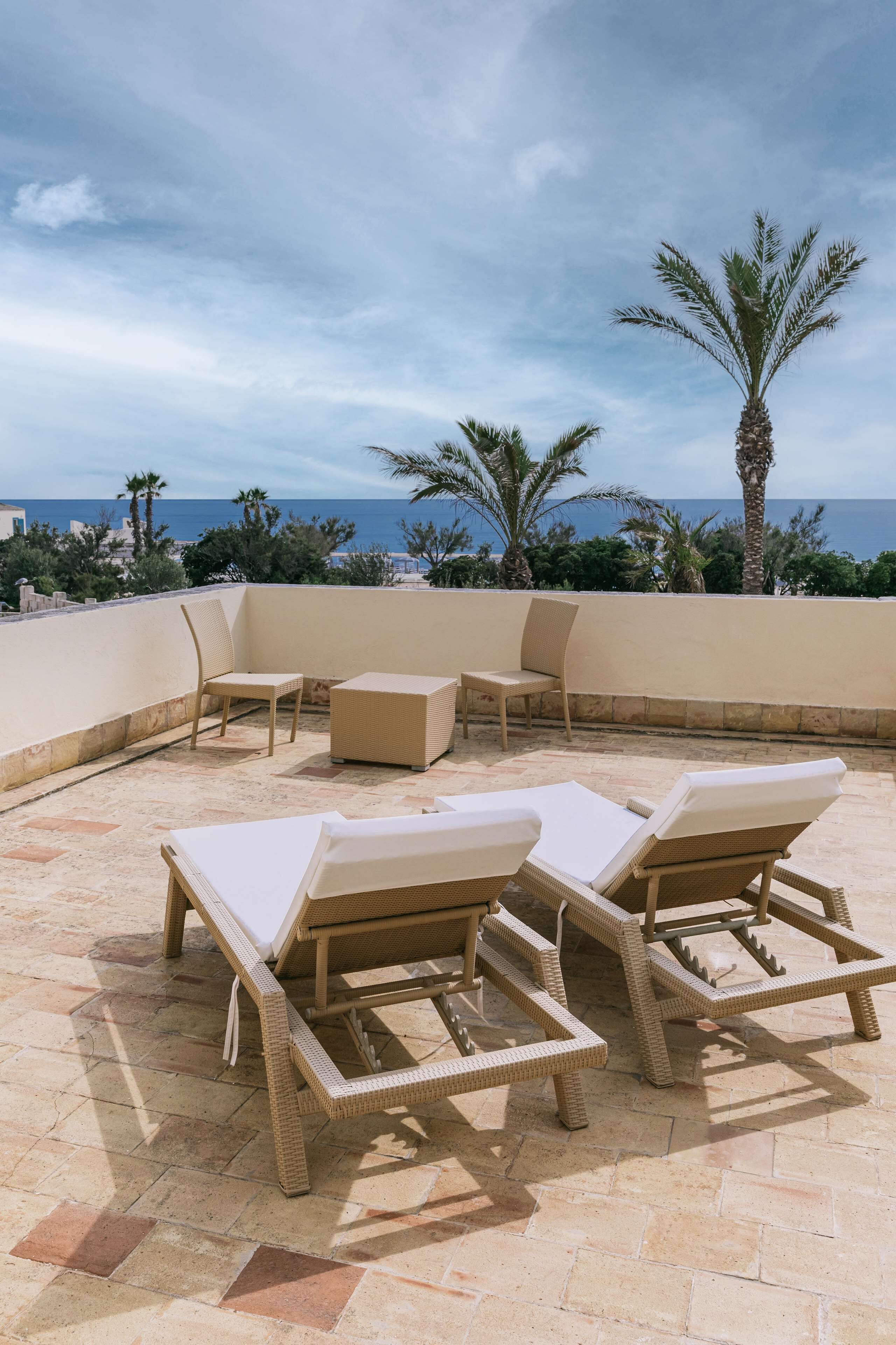 A rooftop terrace with wicker lounge chairs and a small table, overlooking a coastal area with palm trees and the sea.