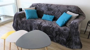 Ein graues, gepolstertes Sofa mit drei blauen Kissen, ein kleiner grauer Tisch und ein gelber Tisch.