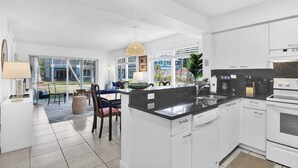A modern kitchen with white cabinets, a black countertop, and a large island. There is a dining area with a table and chairs, and a view of the outside through large windows. - 260B - Boca Retreat w/ Pool, Parking, Near Evert Tennis & Shops (Boca Raton)