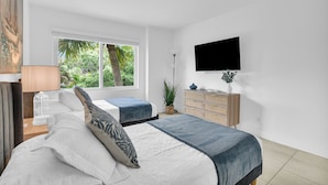 A modern bedroom with a large bed, a TV, a window with a view of greenery, and a bedside table with a lamp. - 260C - Apt in Boca Raton- Near Evert & Boca Prep (Boca Raton)