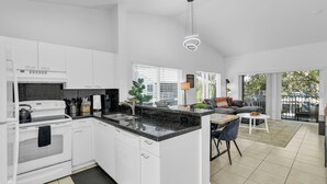 A modern kitchen with white cabinets, a black countertop, and a built-in oven. There is a dining area with a white table and chairs, and a sofa with a view of the outdoors. - 260C - Apt in Boca Raton- Near Evert & Boca Prep (Boca Raton)