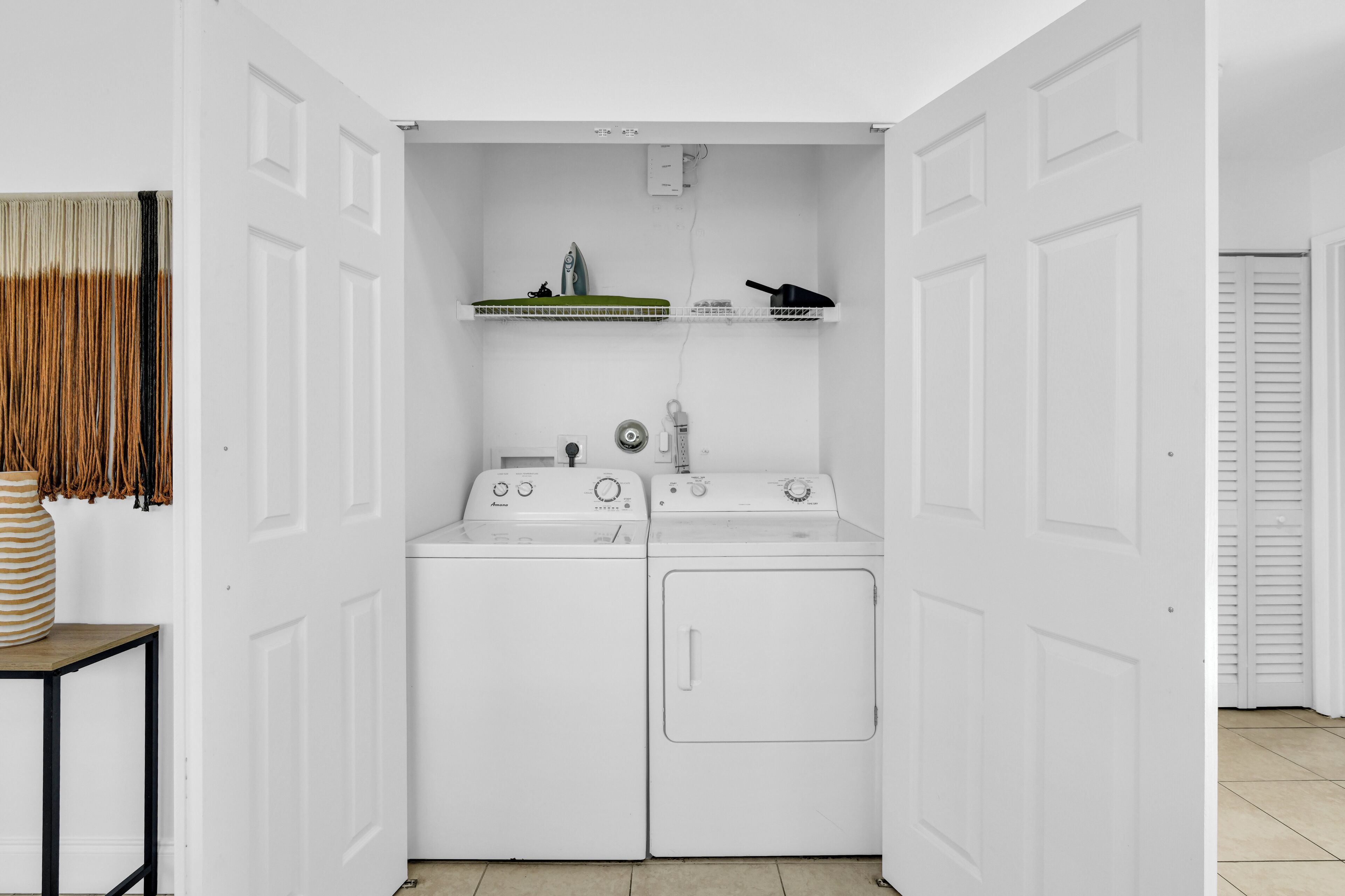 A compact laundry room with two washing machines and a dryer, a small table with a vase, and a shelf with a hairdryer and other items.