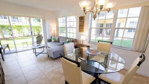 A spacious living area with a glass dining table, comfortable seating, and large windows offering a view of the outdoors. - 270B - Boca Retreat w/ Pool, Parking, Near Evert Tennis & Shops (Boca Raton)
