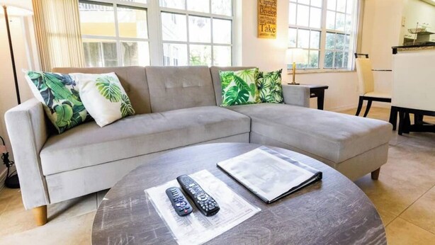 A modern living room with a grey sectional sofa, a round coffee table, and a dining area with a white table and chairs. - 270B - Boca Retreat w/ Pool, Parking, Near Evert Tennis & Shops (Boca Raton)