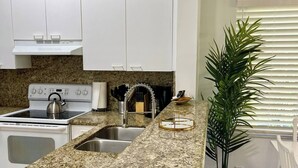 Una cucina con mobili bianchi, un piano di lavoro in granito, un fornello, un lavello e una pianta in vaso.