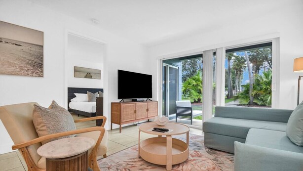 A modern living room with a sofa, armchair, and coffee table. A television is mounted on the wall, and there is a sliding glass door leading to a balcony with plants. - 350B - Boca Retreat w/ Pool, Parking, Near Evert Tennis & Shops (Boca Raton)