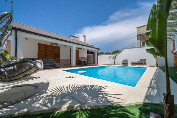 A pool area with a clear blue pool, a covered seating area, and a patio with a table and chairs.