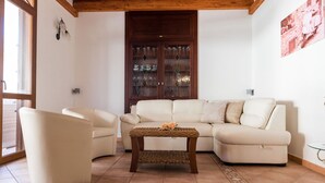 A living room with a beige sofa, two armchairs, a coffee table with a woven top, a wooden cabinet with glass doors, and a window with a view of a scooter.