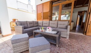 A cozy outdoor seating area with a grey sofa, wicker ottomans, and a coffee table set with pastries and flowers.