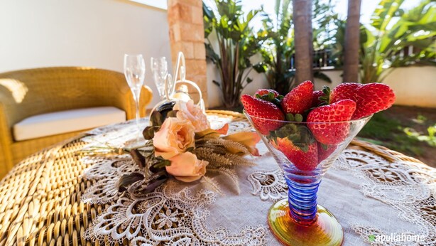 A table with a glass vase containing strawberries, a lace tablecloth, and a wicker chair in the background.