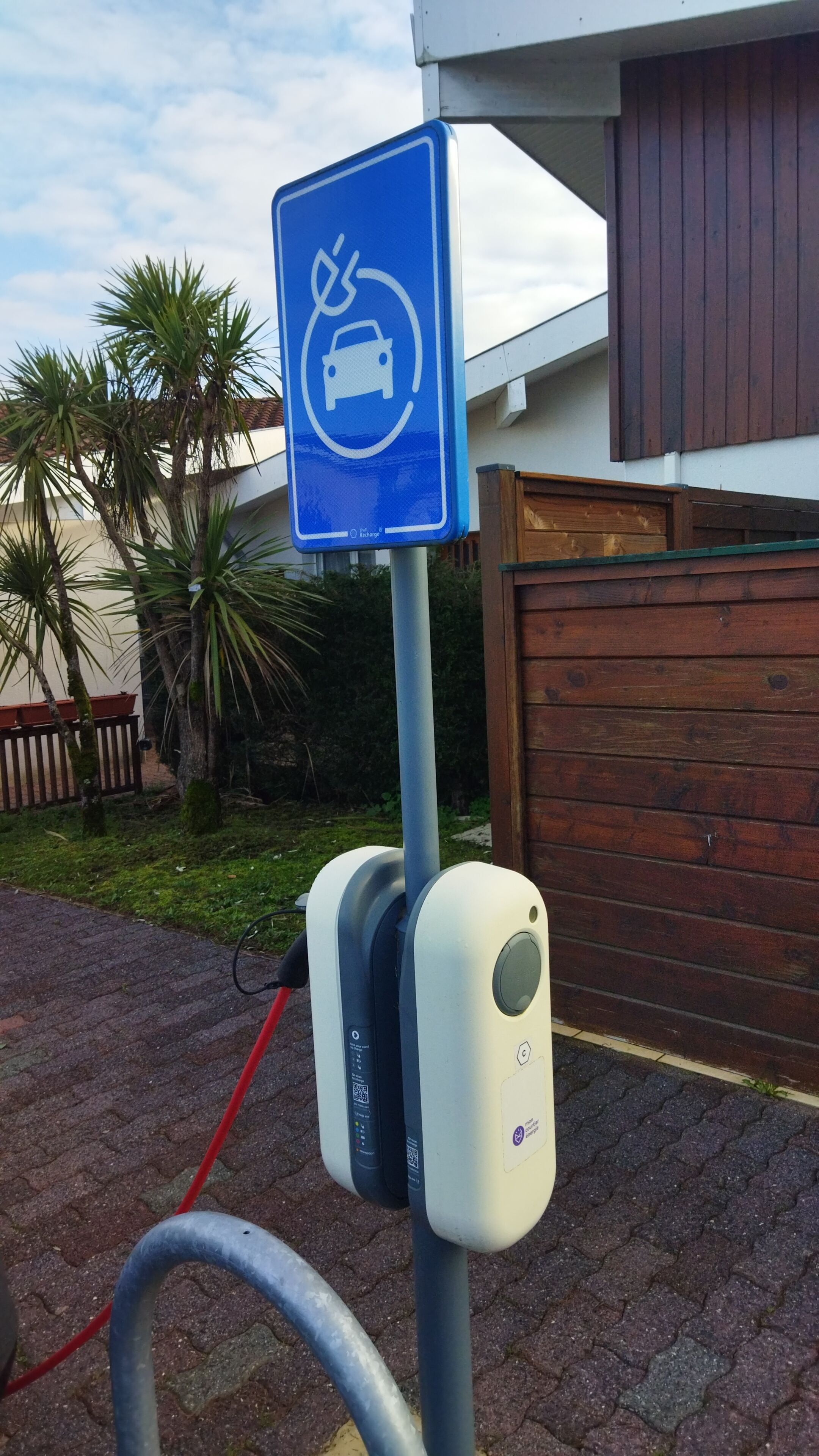 A parking sign with a car and a prohibition symbol, a charging station, and a wooden fence in the background.