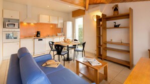 A modern kitchen with white cabinets, a dining area with a blue sofa and wooden table, and a bookshelf with decorative items. - Résidence Amaréna (Saint-Paul-les-Dax)
