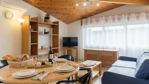 A cozy living room with a wooden ceiling, a dining table set with plates, glasses, and a bottle, a sofa, and a TV on a wooden stand. - Résidence Amaréna (Saint-Paul-les-Dax)
