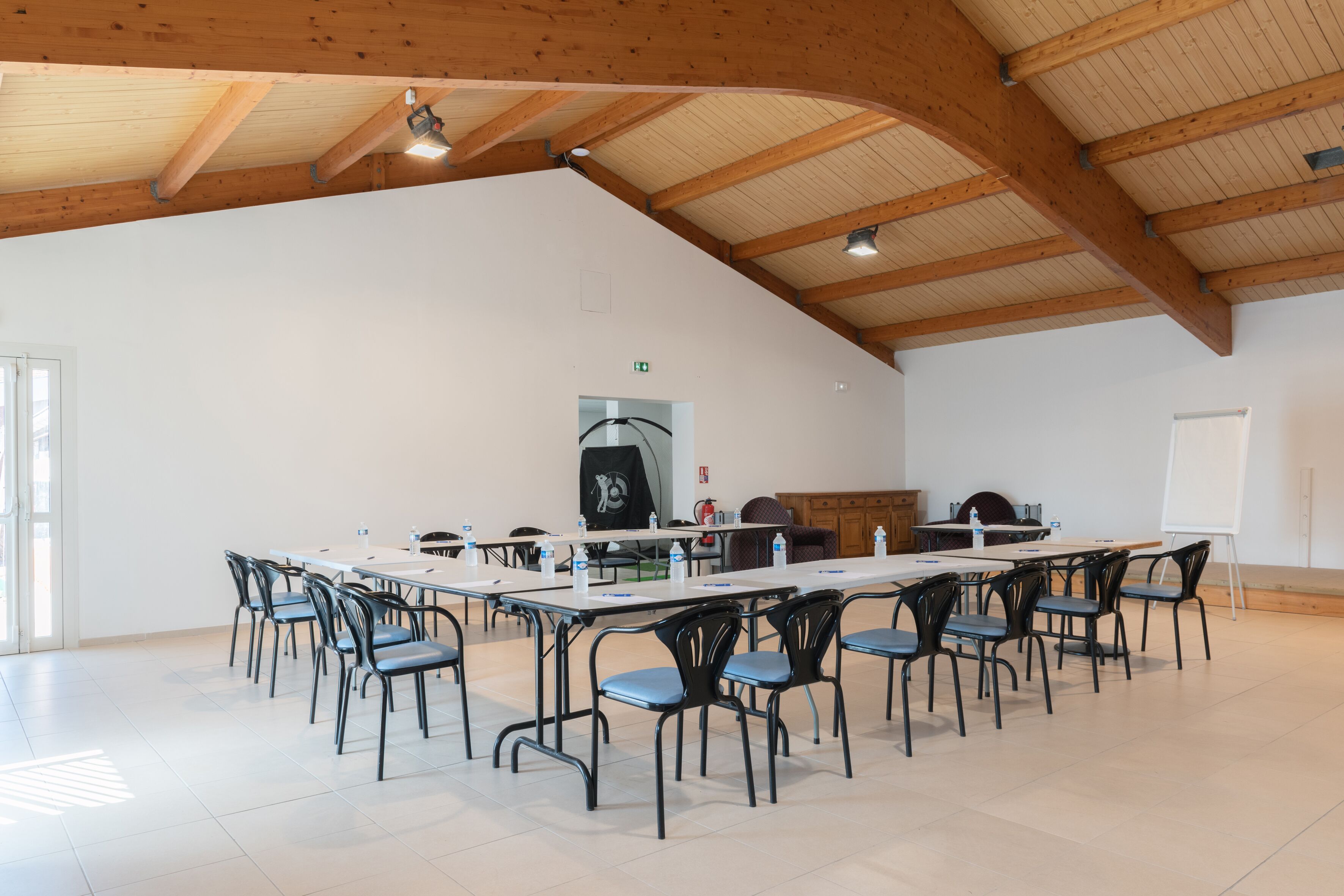 Una habitación amplia con techo alto y vigas de madera, que cuenta con una mesa larga con sillas, una pizarra y un dispensador de agua.