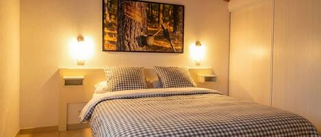 A bedroom with a bed, checkered bedding, two bedside lamps, a wall-mounted painting, and a wooden ceiling.