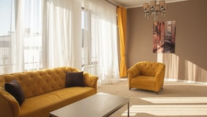 Una sala de estar con un sofá amarillo, un sillón a juego, una mesa de centro, cortinas translúcidas y una araña.