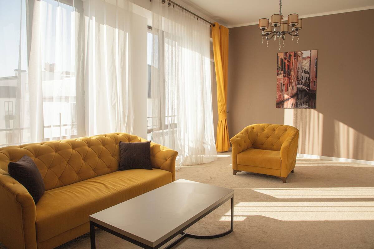 A living room with a yellow sofa, a small armchair, a coffee table, and sheer curtains.