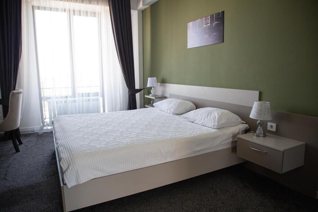 Una cama bien hecha con sábanas blancas, una mesita de noche con lámpara, y una silla en una habitación con paredes verdes y un ventanal grande.