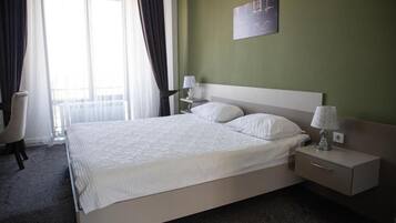 Una cama bien hecha con sábanas blancas, una mesita de noche con lámpara, y una silla en una habitación con paredes verdes y un ventanal grande.