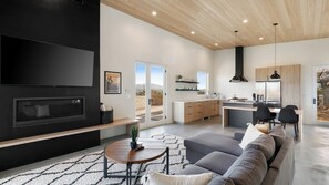A modern living room with a flat-screen TV, a fireplace, a sofa, and a dining area with a kitchen in the background.