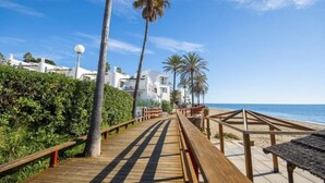 A wooden boardwalk leading to a beach with palm trees and white buildings in the background.