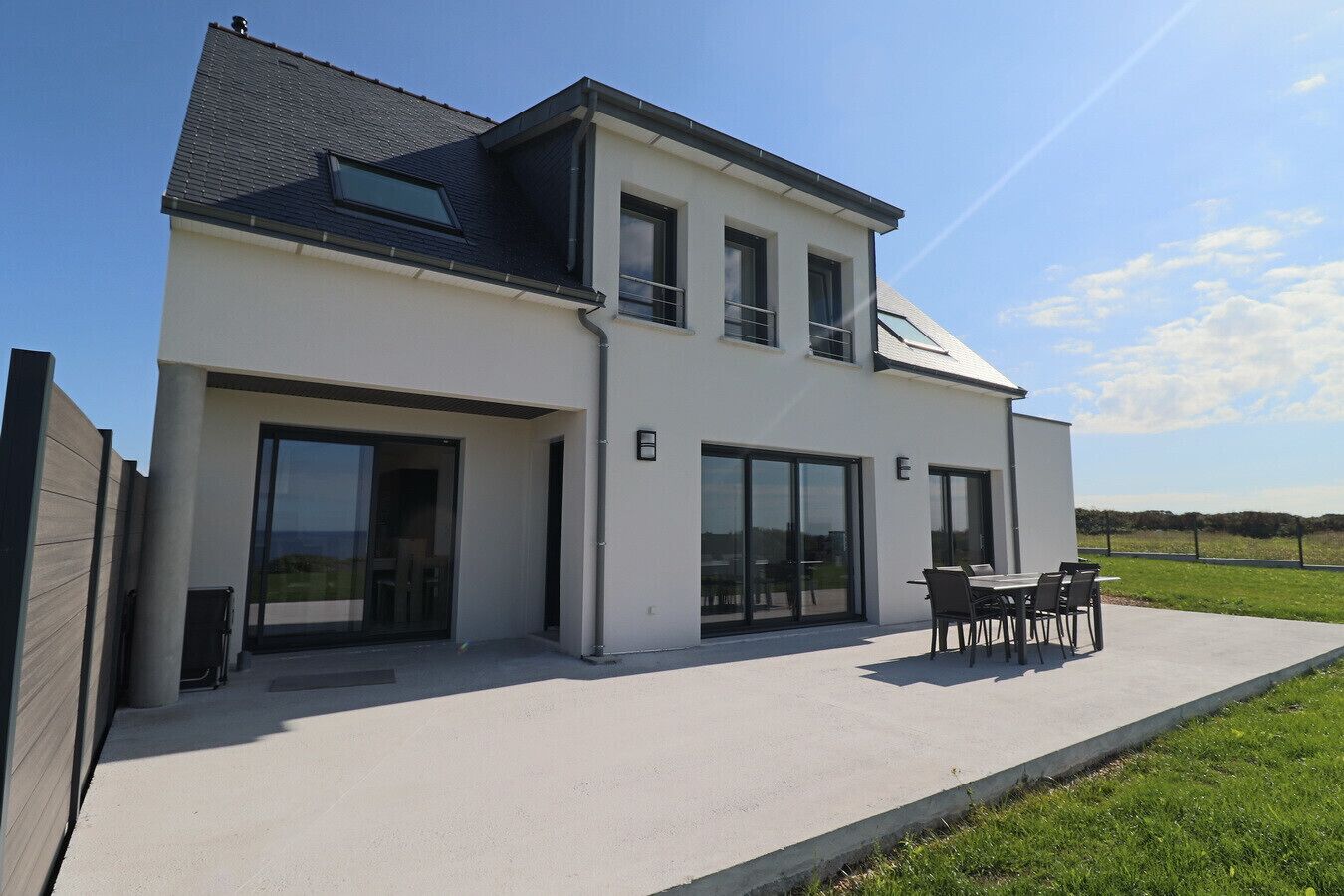 A modern two-story house with large sliding glass doors, a patio with outdoor furniture, and a view of the countryside.