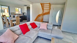Ein modernes Wohnzimmer mit einem grauen Ecksofa, roten und pinkfarbenen Kissen, einer Holztreppe und einem Essbereich im Hintergrund.