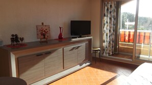 A room with a wooden cabinet, a flat-screen TV, a painting, and a view of the outdoors through a sliding glass door. - Studio rental (Saint-Mandrier-sur-Mer)