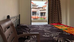 Une chambre avec un lit, une chaise, une couvre-lit à motifs, un rideau et une vue sur un immeuble doté de balcons.