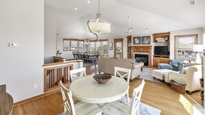 A spacious living area with a round dining table, white chairs, a beige sofa, and a television in the background. - CheckMate: Walk to Beach, Large Pool, Hot Tub (Southern Shores)