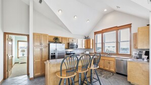 A modern kitchen with wooden cabinets, a stainless steel refrigerator, a built-in oven, and a large window providing natural light. - CheckMate: Walk to Beach, Large Pool, Hot Tub (Southern Shores)