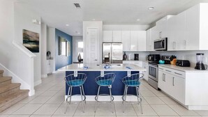 A modern kitchen with white cabinets, a central island with bar stools, stainless steel appliances, and a staircase leading to another level. - Entertaining Retreat with Themed Bedrooms at Encore (Kissimmee)