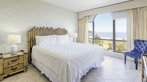A hotel room with a large bed, two bedside tables with lamps, a view of the ocean, and a blue chair.