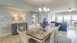 A dining area with a glass-top table, white chairs, a blue sofa, and a view of the ocean through large windows.