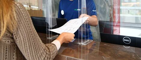A reception desk with a person behind a transparent barrier, a computer monitor, and a menu stand.