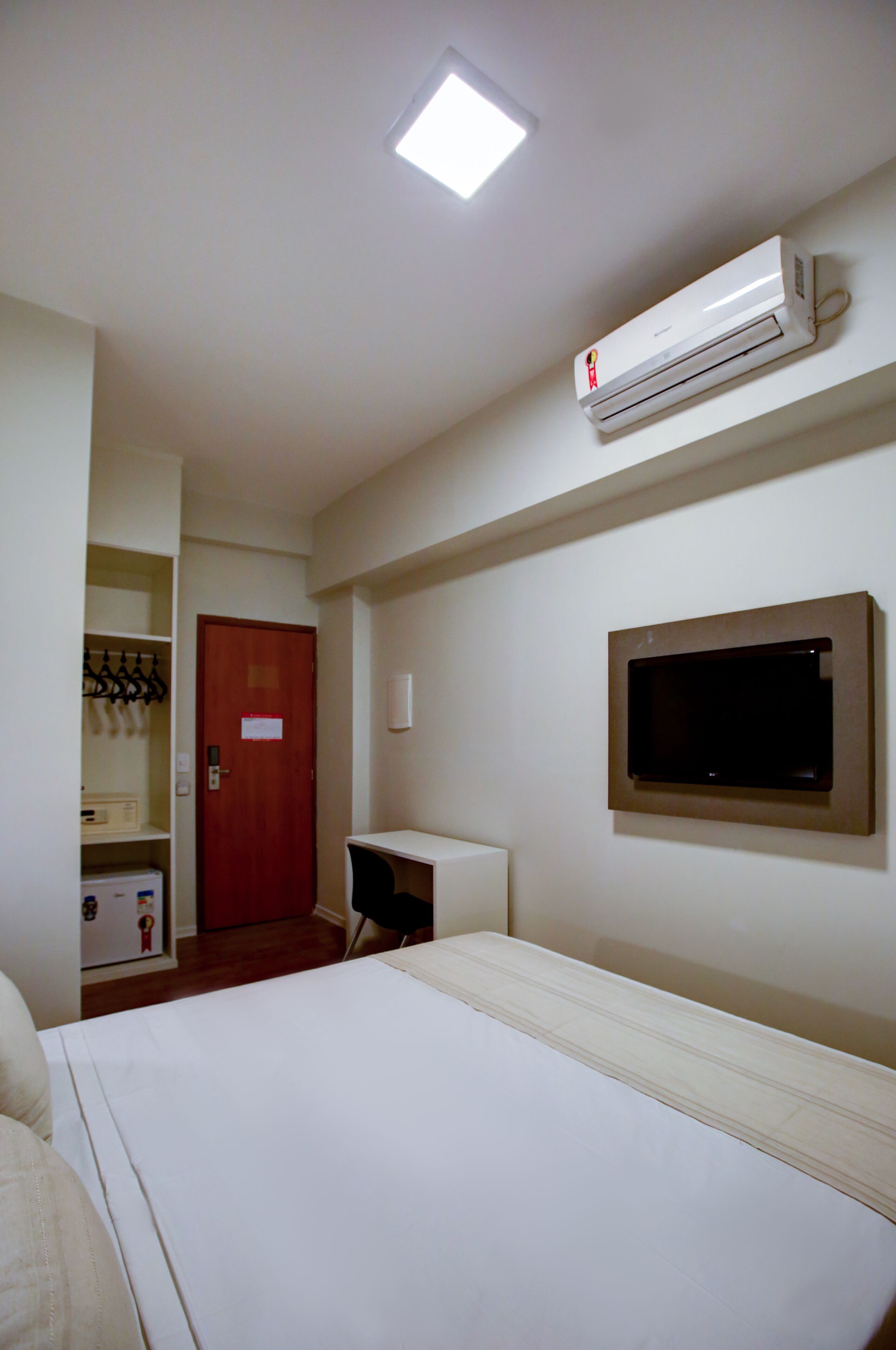 A hotel room with a bed, a desk, a TV mounted on the wall, an air conditioner, a coat rack, and a door.