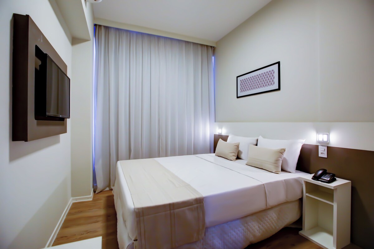 A hotel room with a bed, a bedside table with a phone, a wall-mounted TV, and a window with curtains.